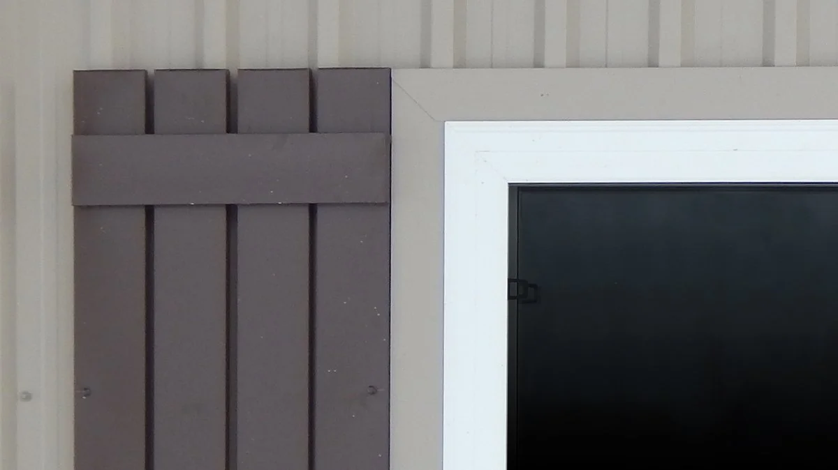 Close-up of a partial view of a house exterior showing a section of a gray fence or panel to the left and a window with a white frame to the right.