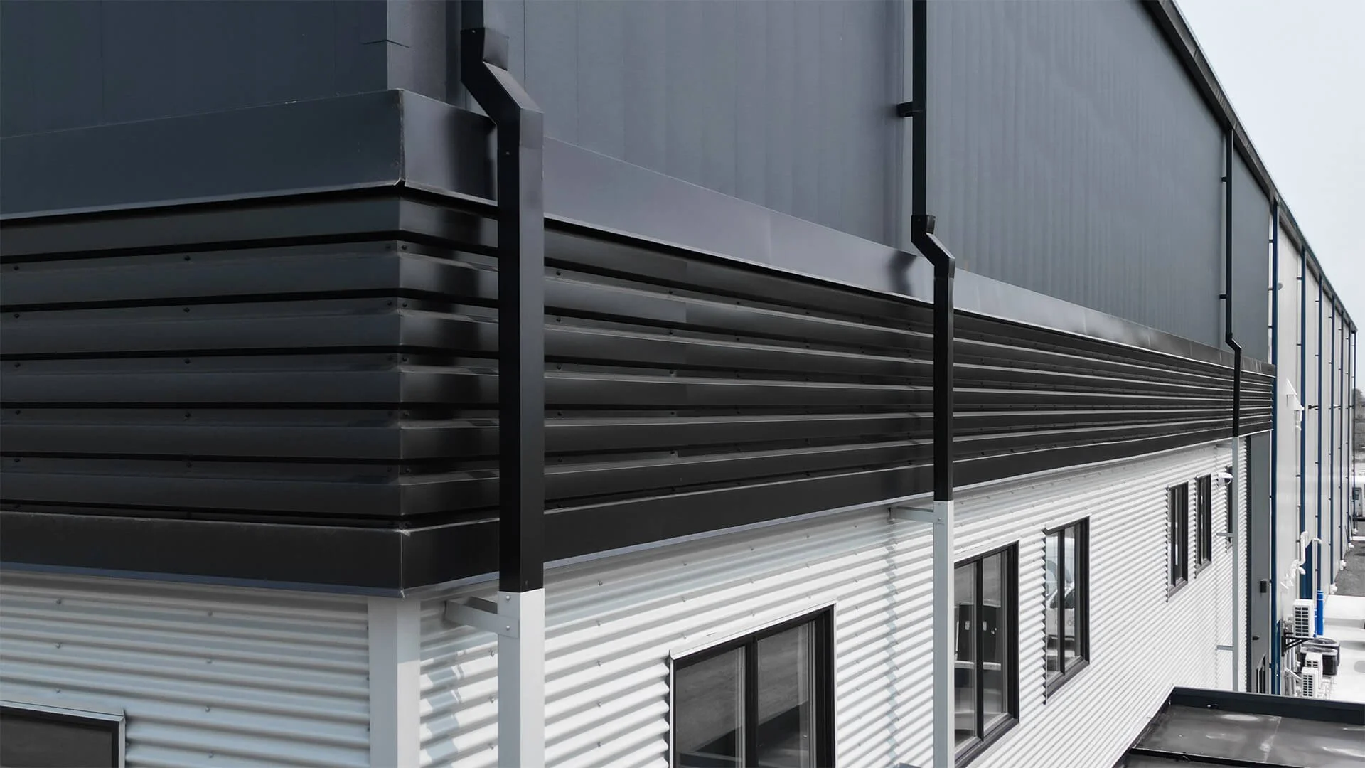 Exterior of a modern industrial building with black and white metal siding, multiple windows, and external heating or air conditioning units.