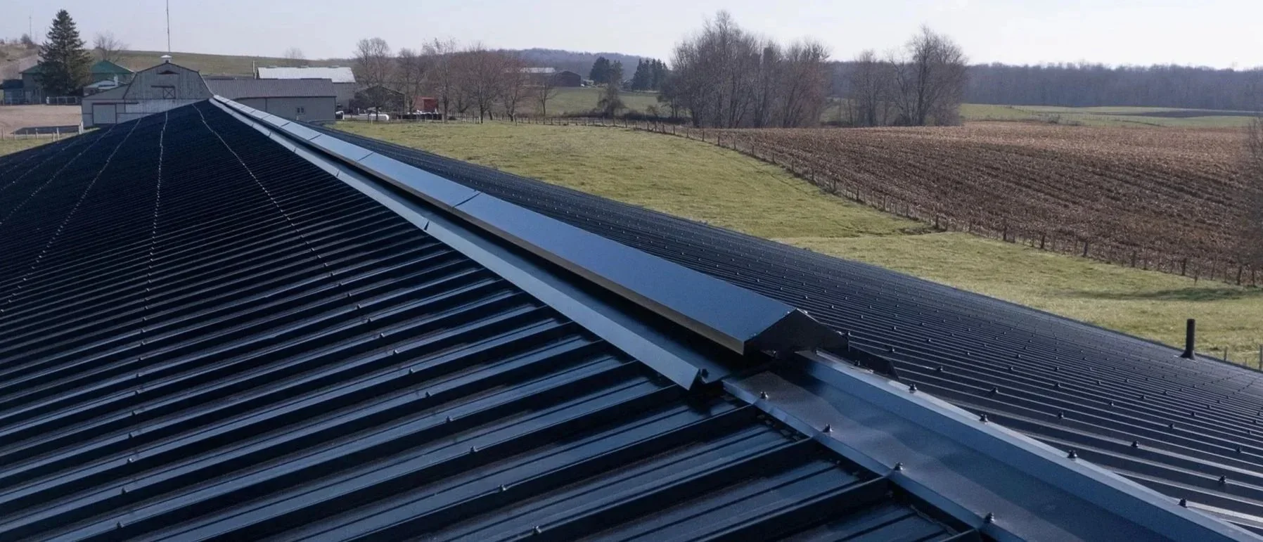 Close-up view of a black metal roof with solar panels installed on top, overlooking a rural landscape with fields, trees, and farm buildings in the background.