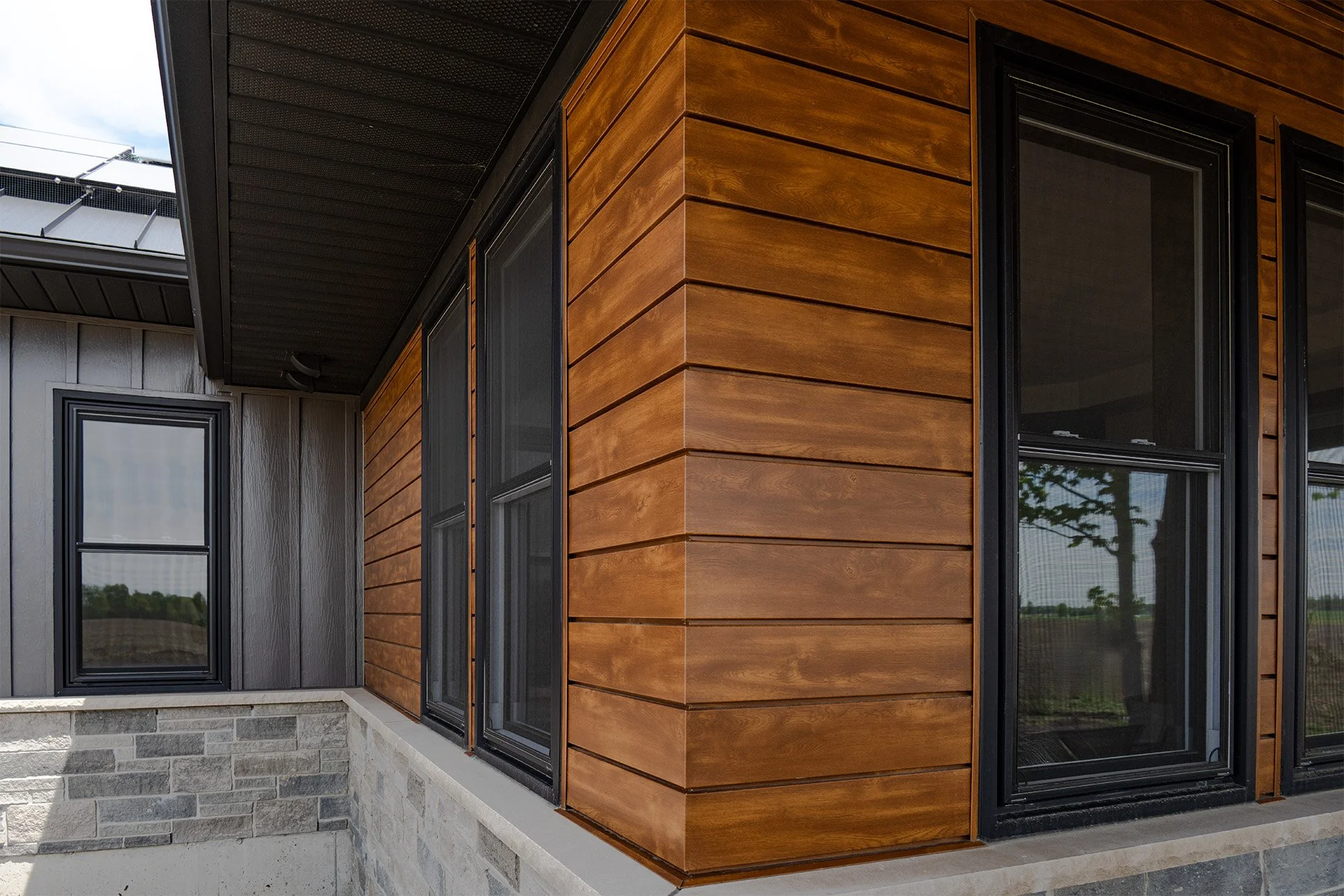 Close-up of a modern house corner with wooden siding, black-framed windows, and a stone foundation.
