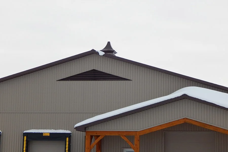 A gray metal building with snow on the roof, featuring a gable and an attic vent in the upper section, and a smaller front extension with an overhang, also covered in snow.