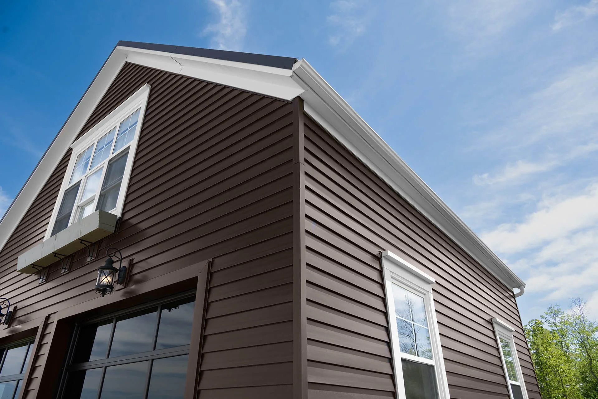 Close-up view of a brown wooden house with white-framed windows and black lantern-style exterior lights under a blue sky with wispy clouds.