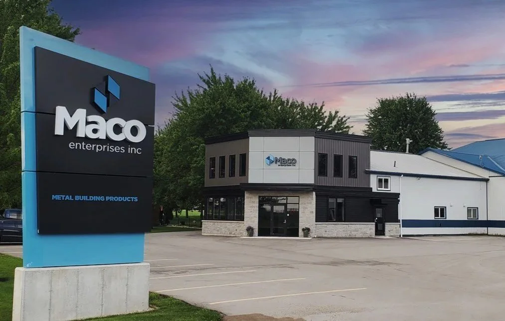 The image shows the building and parking lot of Maco Enterprises Inc., a company that produces metal building products, with a large sign in the foreground and sunset sky in the background.
