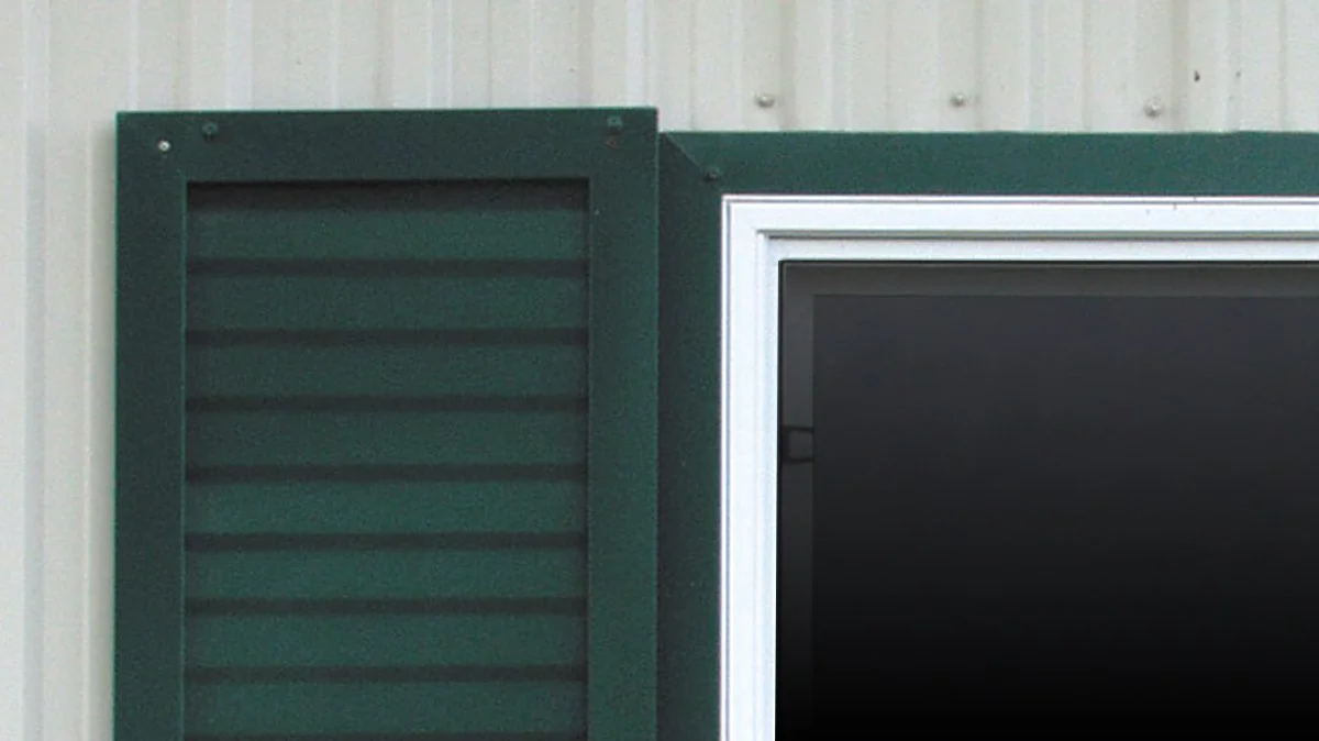 Close-up of green louvered window shutter next to a black window with a silver frame outside a beige wall.