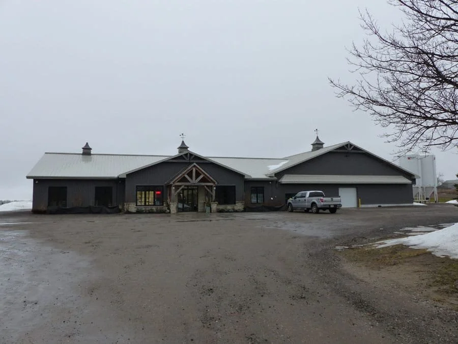 An exterior view of a modern barn-style building with a gray metal roof and dark gray walls, with a parking area in front and snow patches on the ground.