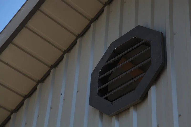A black octagonal vent with horizontal slats mounted on a metal building with vertical siding.