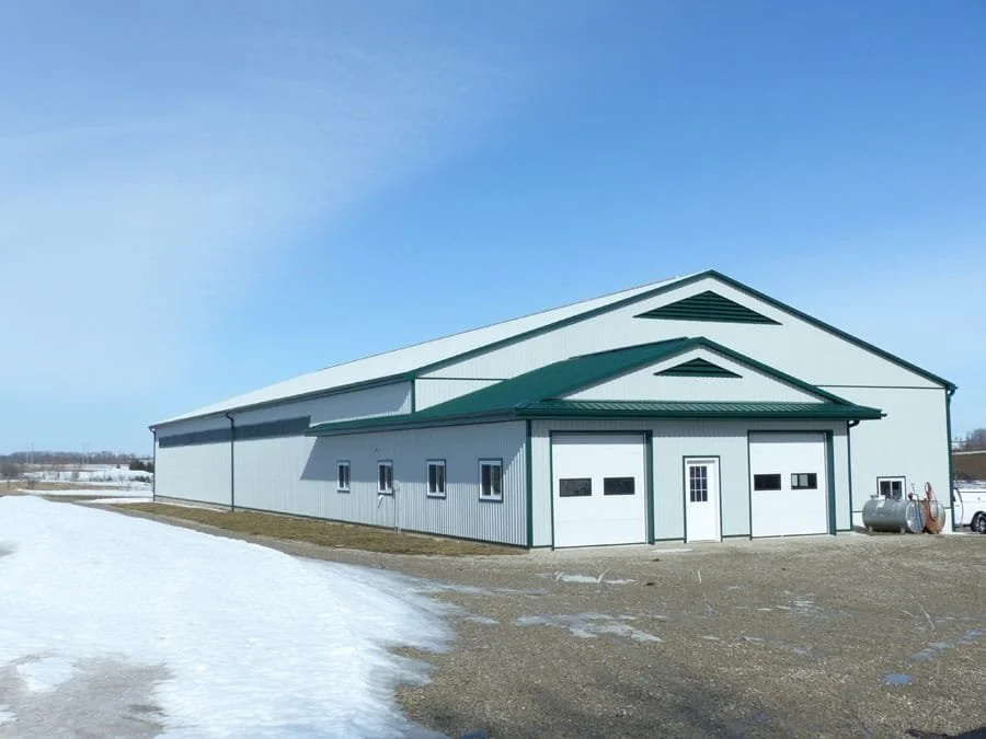 A large metal storage building with multiple garage doors, a small door, and small windows, located in a rural area with snow on the ground and a clear blue sky.