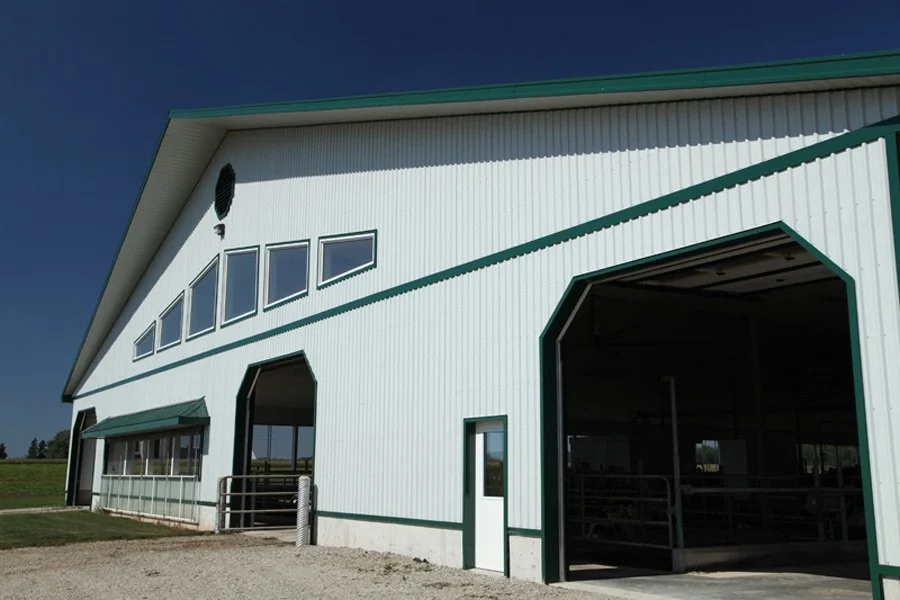 A large white metal barn with green trim and an open entryway, some small windows, and a clear blue sky.