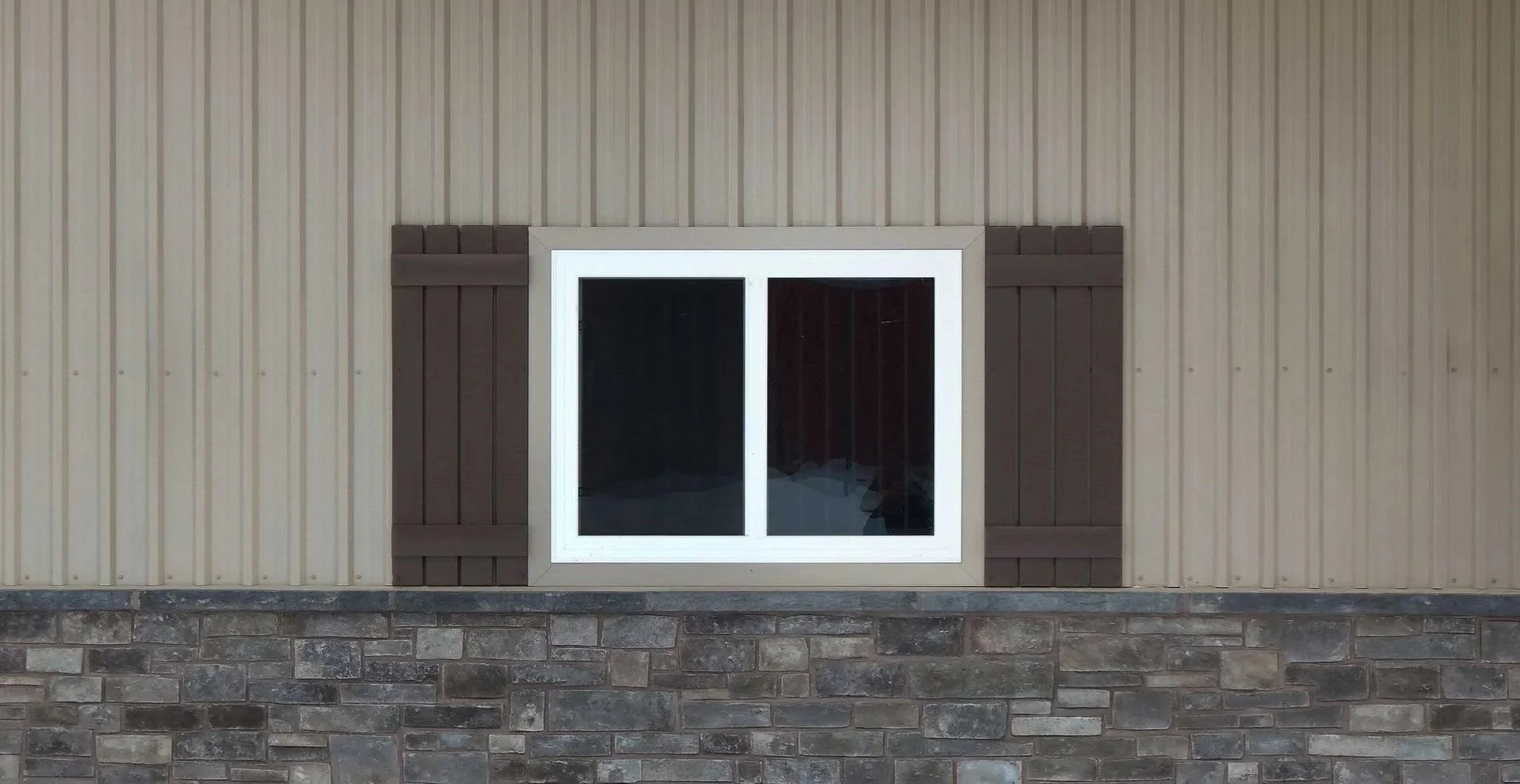 A window with a white frame on a beige vertical siding wall, with decorative brown wood panels on each side, and a stone foundation below.