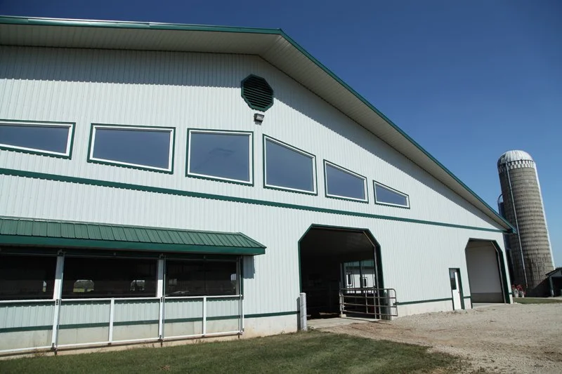 Large white farm barn with green trim, multiple windows, and a silo in the background under a clear blue sky.