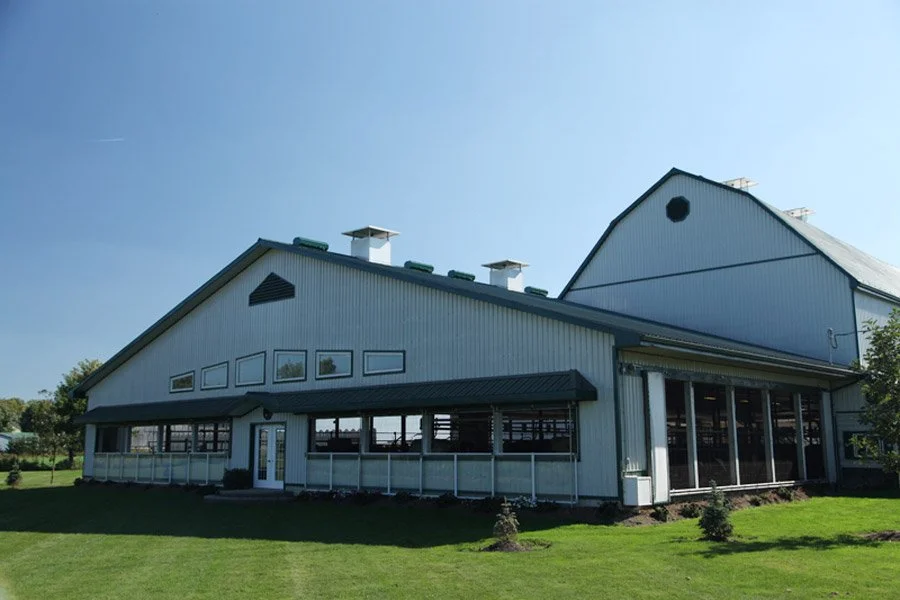 A large, modern barn with white walls, a gray roof, and several ventilation structures on top, set on a green lawn under a clear blue sky.