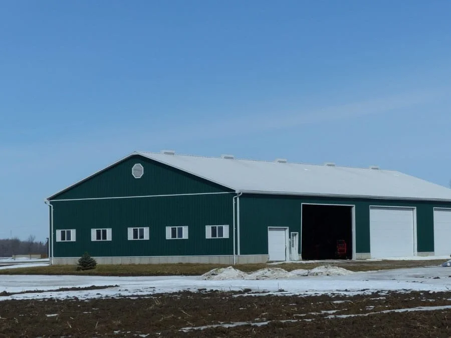 A large green barn with a white roof, partially open garage door, and small windows, set in a rural landscape with patches of snow on the ground and a clear blue sky.