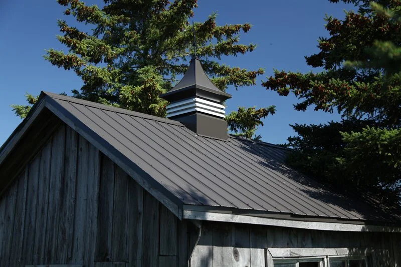 A close-up view of a metal roof on a wooden building with a decorative chimney cap on top, surrounded by green pine trees against a clear blue sky.