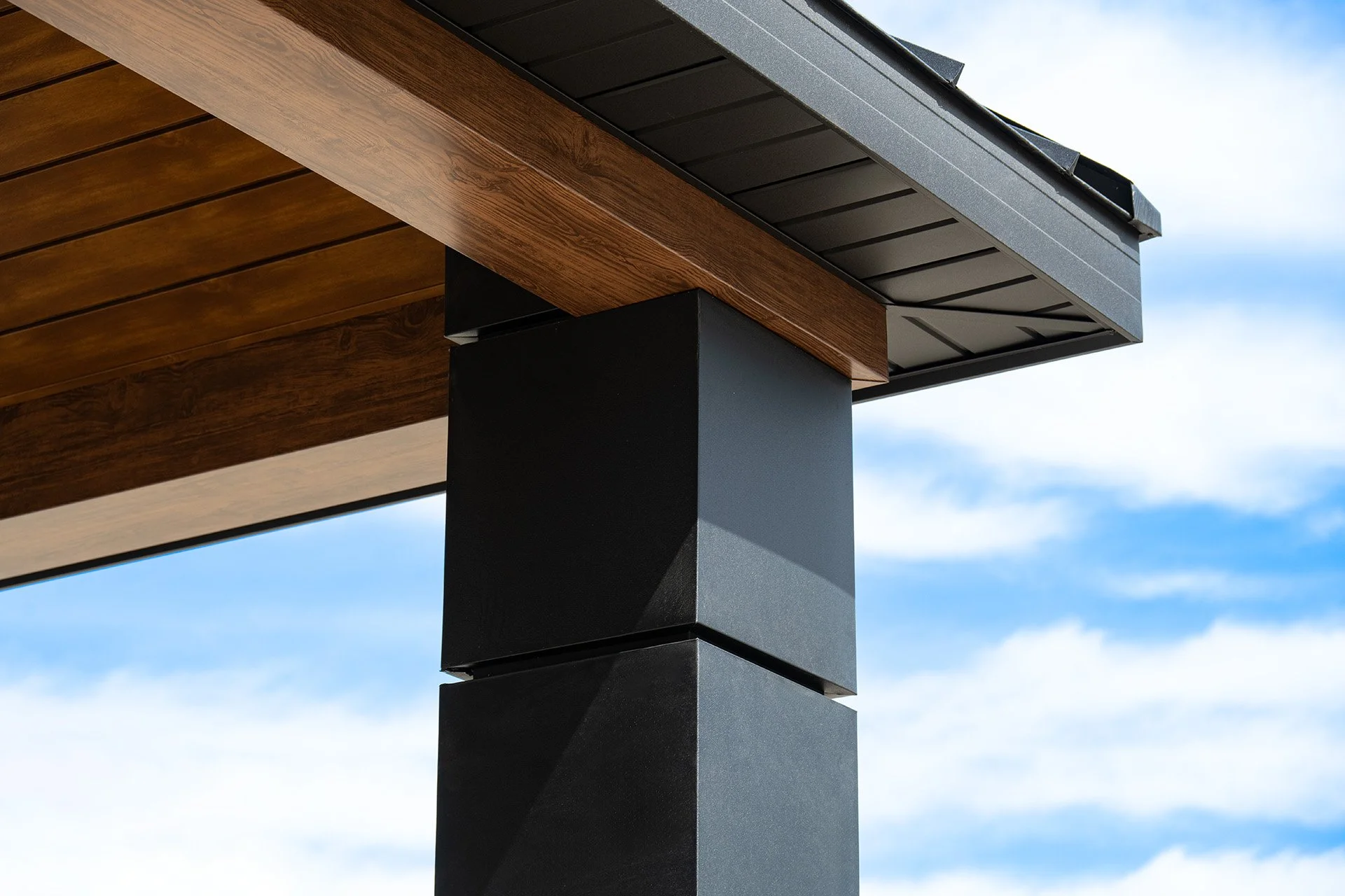 Close-up of a modern building corner with wooden and black metal materials under a blue sky with sparse clouds.