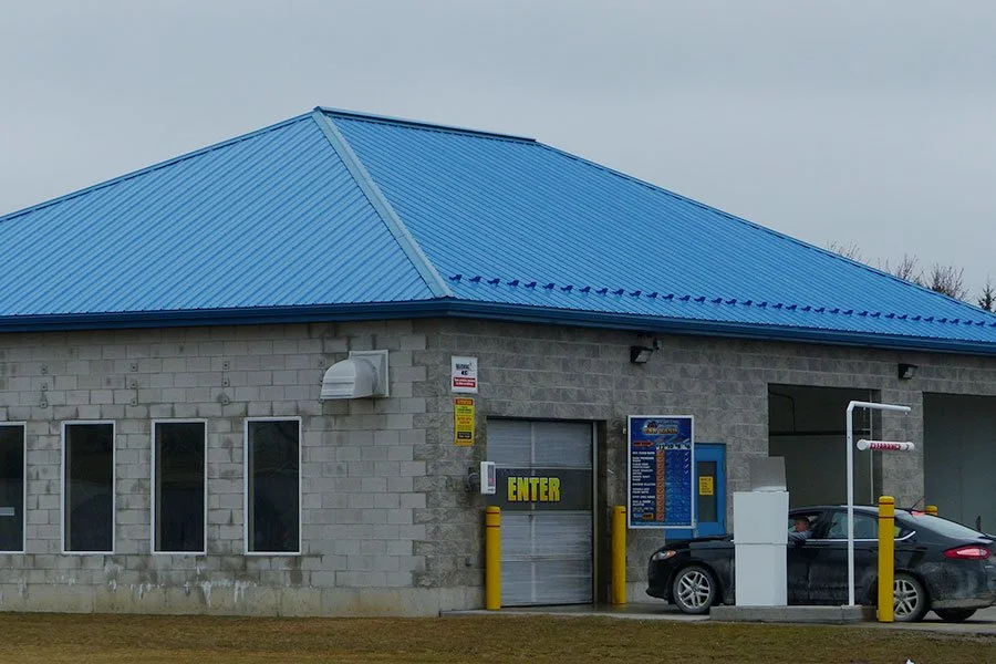 A roadside auto wash station with a blue metal roof, concrete block walls, and a car waiting inside.