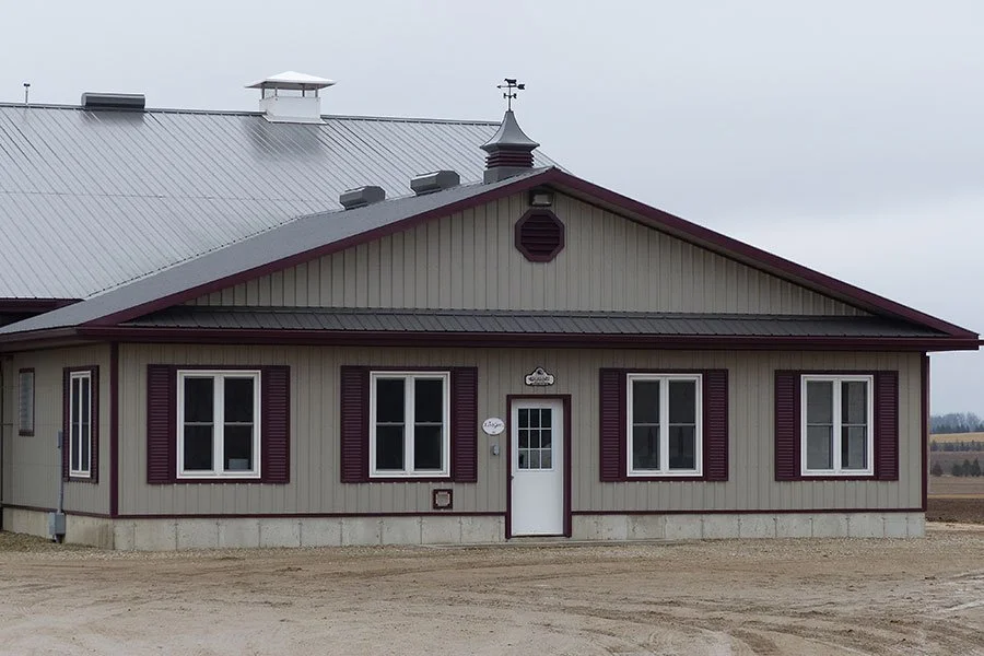A beige house with dark purple shutters, three windows, and a white door, situated on a dirt lot under a cloudy sky.