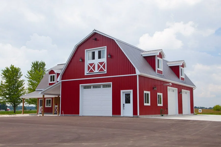Red barn with white accents, two garage doors, and small upper windows, set in a rural area with trees and cloudy sky