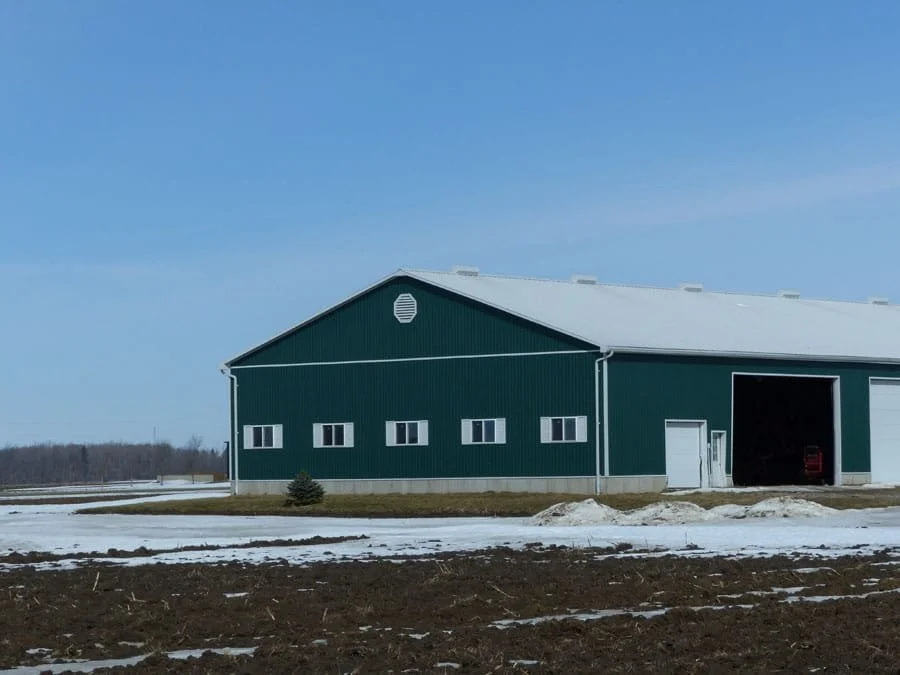 Large green metal barn with a white roof and small windows, located in a rural area with patches of snow on the ground.