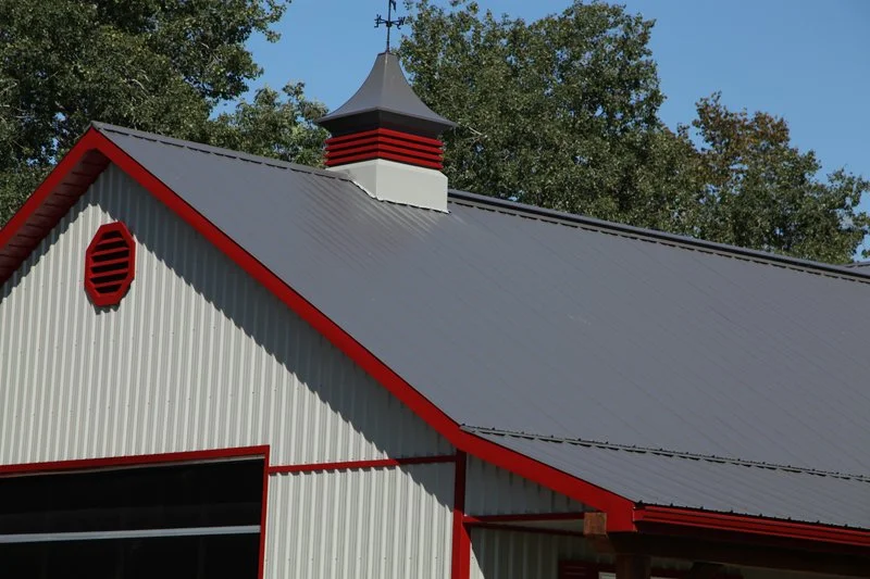 Close-up of a building's roof with a weather vane, painted white and red, under a blue sky with trees in the background.