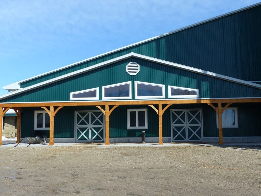 Farm-style barn with green metal siding, white trim, and a large overhang supported by wooden posts. The barn has multiple windows, including triangular and rectangular shapes, and a vent near the roof peak. The ground in front is dirt with a wheelba