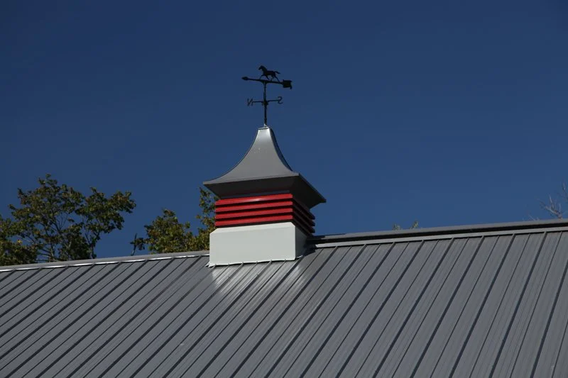 Close-up of a metal roof with a vent topper and weather vane against a clear blue sky.