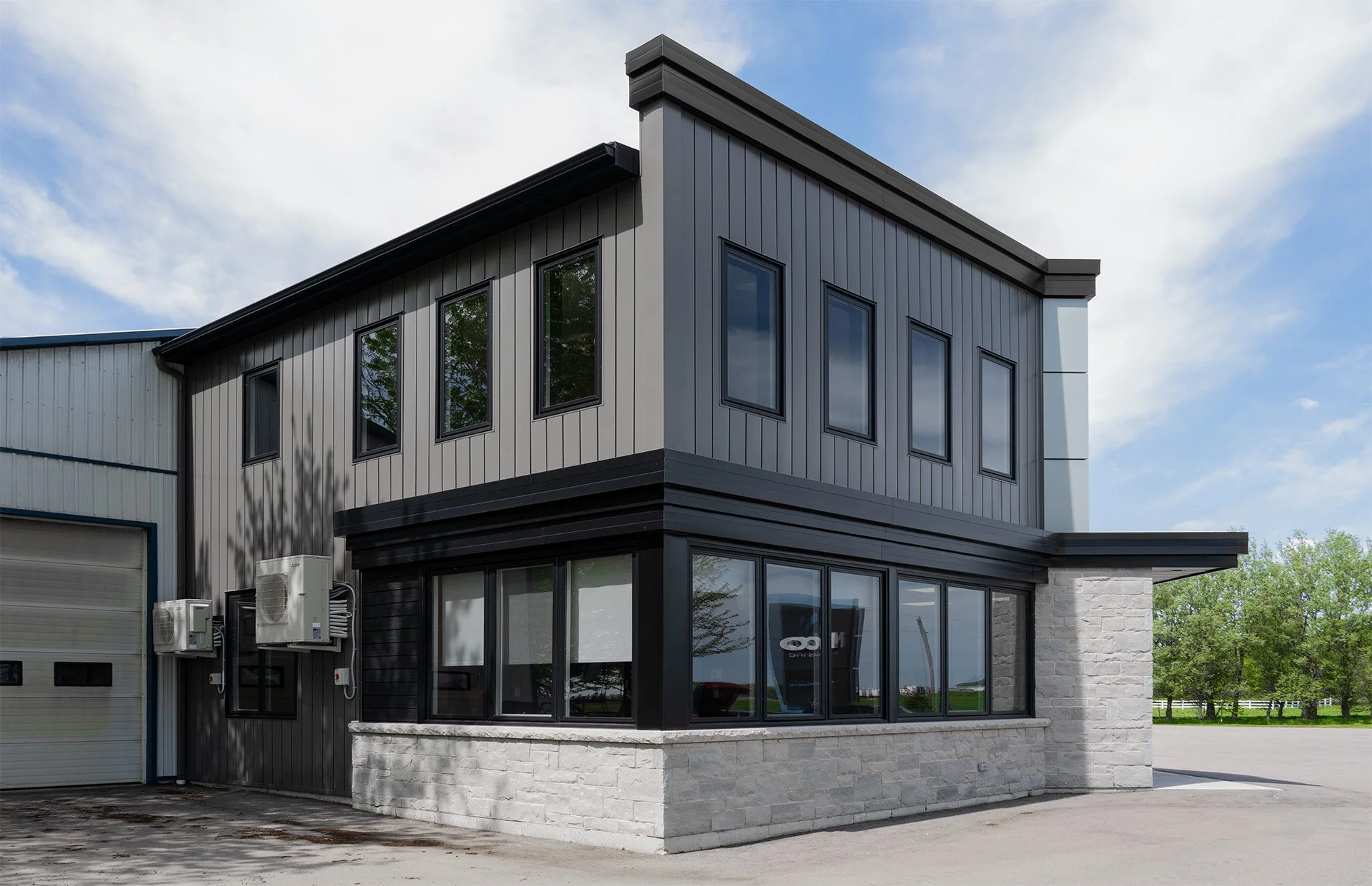 Modern two-story commercial building with dark metal siding, large corner windows on the ground floor, and smaller windows on the upper floor, surrounded by greenery and a clear sky.