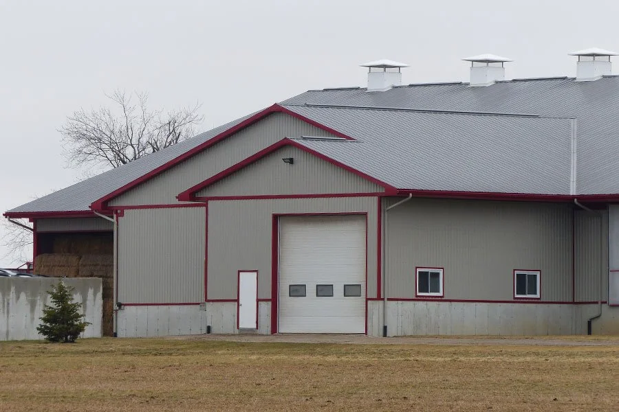 A large, gray barn with red trim, a garage door, small windows, and a small evergreen tree in front.