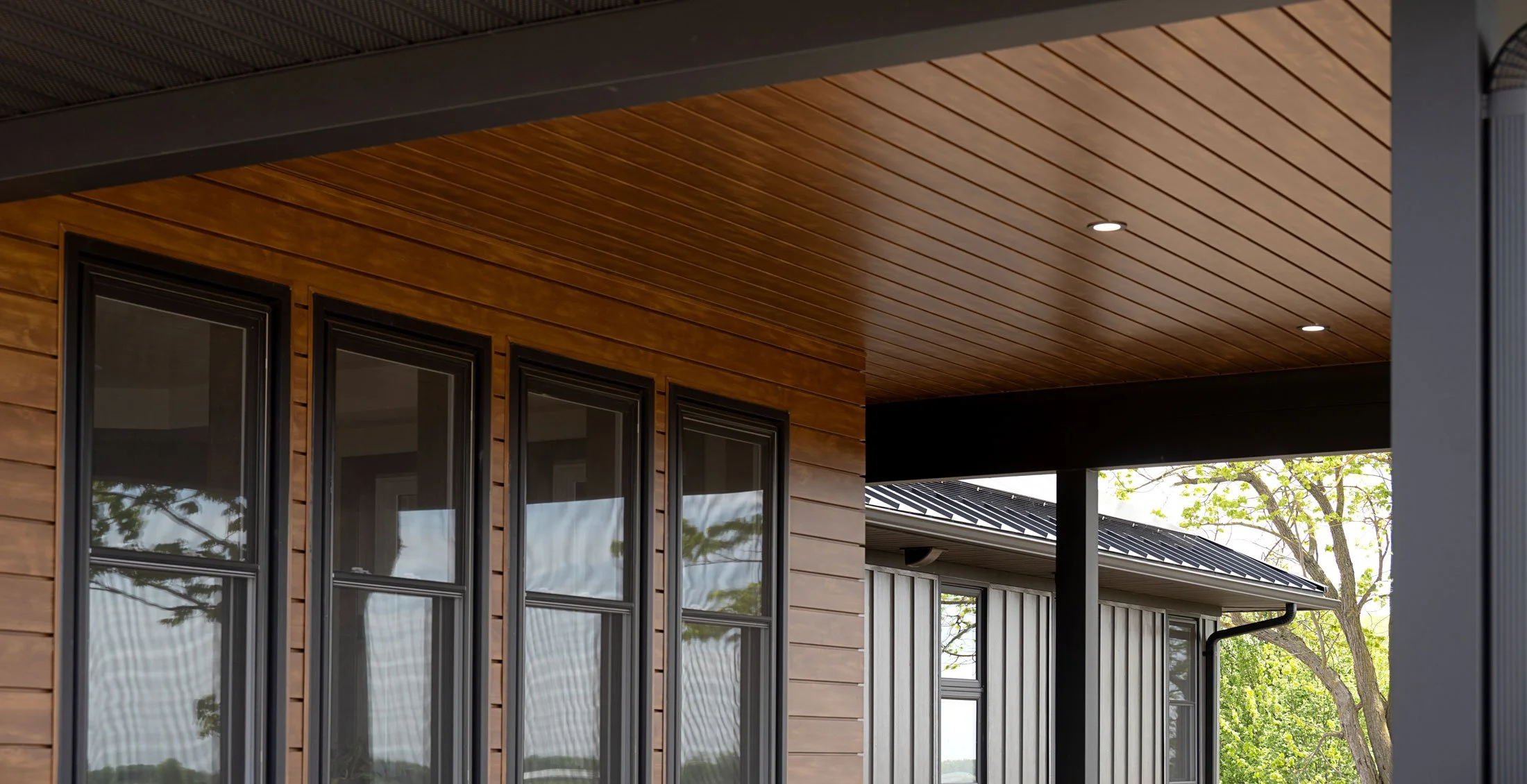 Close-up of modern house exterior with wooden siding, black framed windows, and a covered porch area with recessed lighting, surrounded by trees.