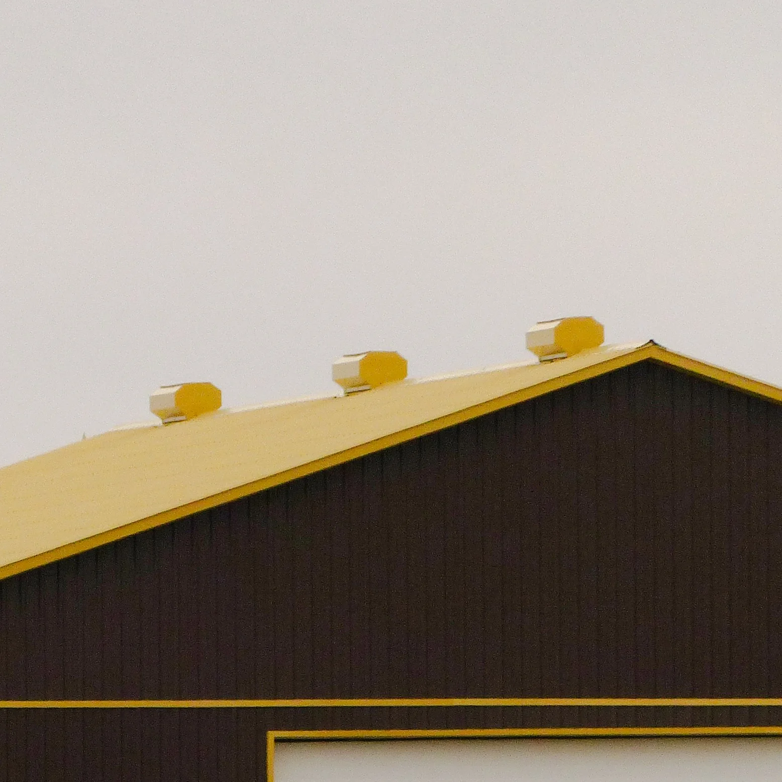 Close-up of a brown building with a yellow roof and three yellow exhaust vents on top, against a cloudy sky.