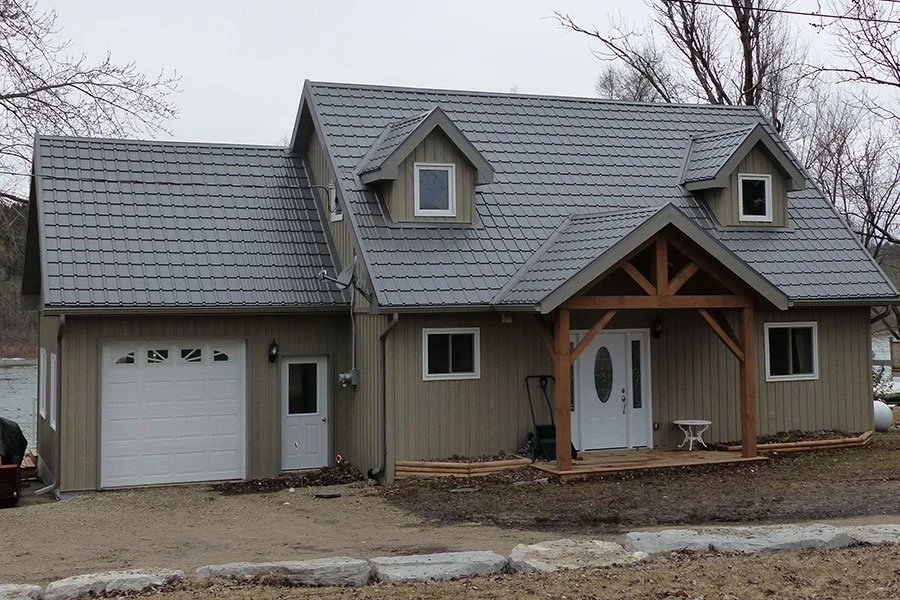 New beige house with gray metal roof, dormer windows, front porch with wooden beams, white front door, and garage, surrounded by dirt and trees in the background.