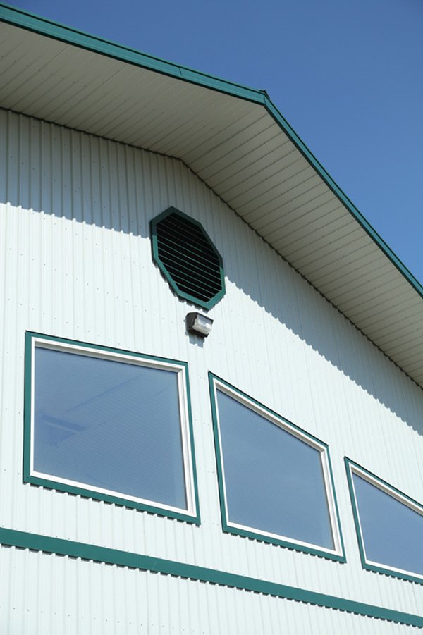 Close-up of the upper exterior of a modern white building with green trim, showing a black vent, a small outdoor light, and three rectangular windows with reflective glass.
