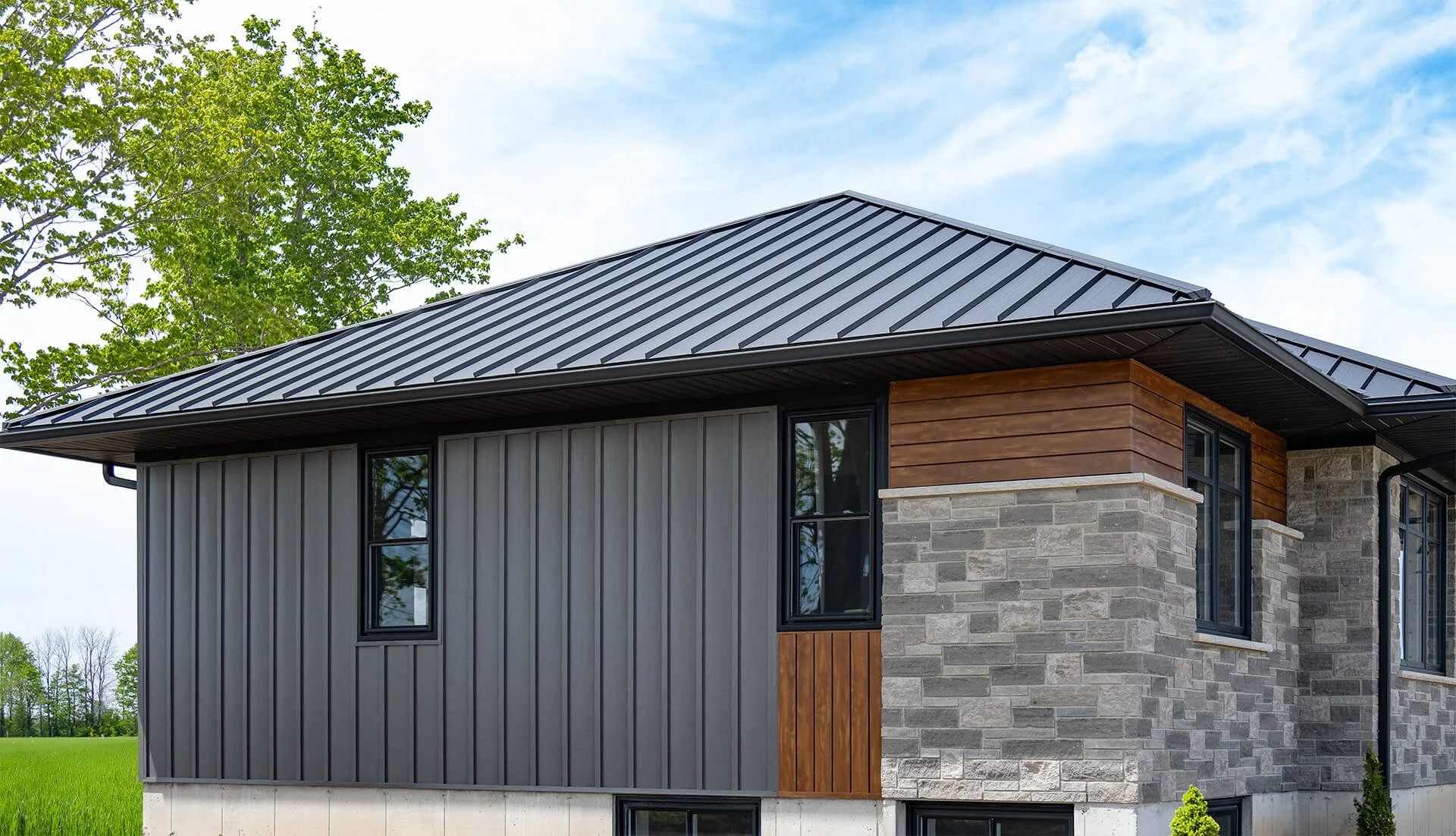 Close-up of a modern house with a metal roof, gray siding, and a stone and wood exterior, surrounded by green grass and trees.