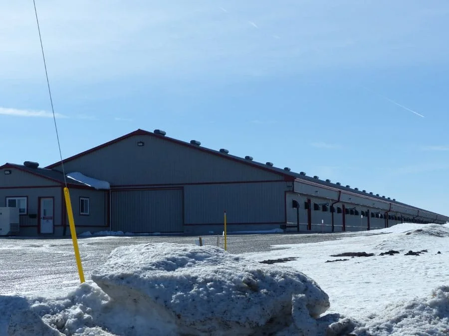 A large industrial building with a red roof and multiple garage doors, surrounded by snow-covered ground and a clear blue sky.