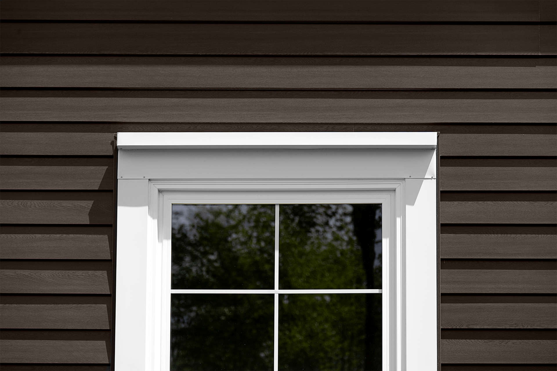 Close-up of a window on a house with brown horizontal siding and a white frame reflecting trees.