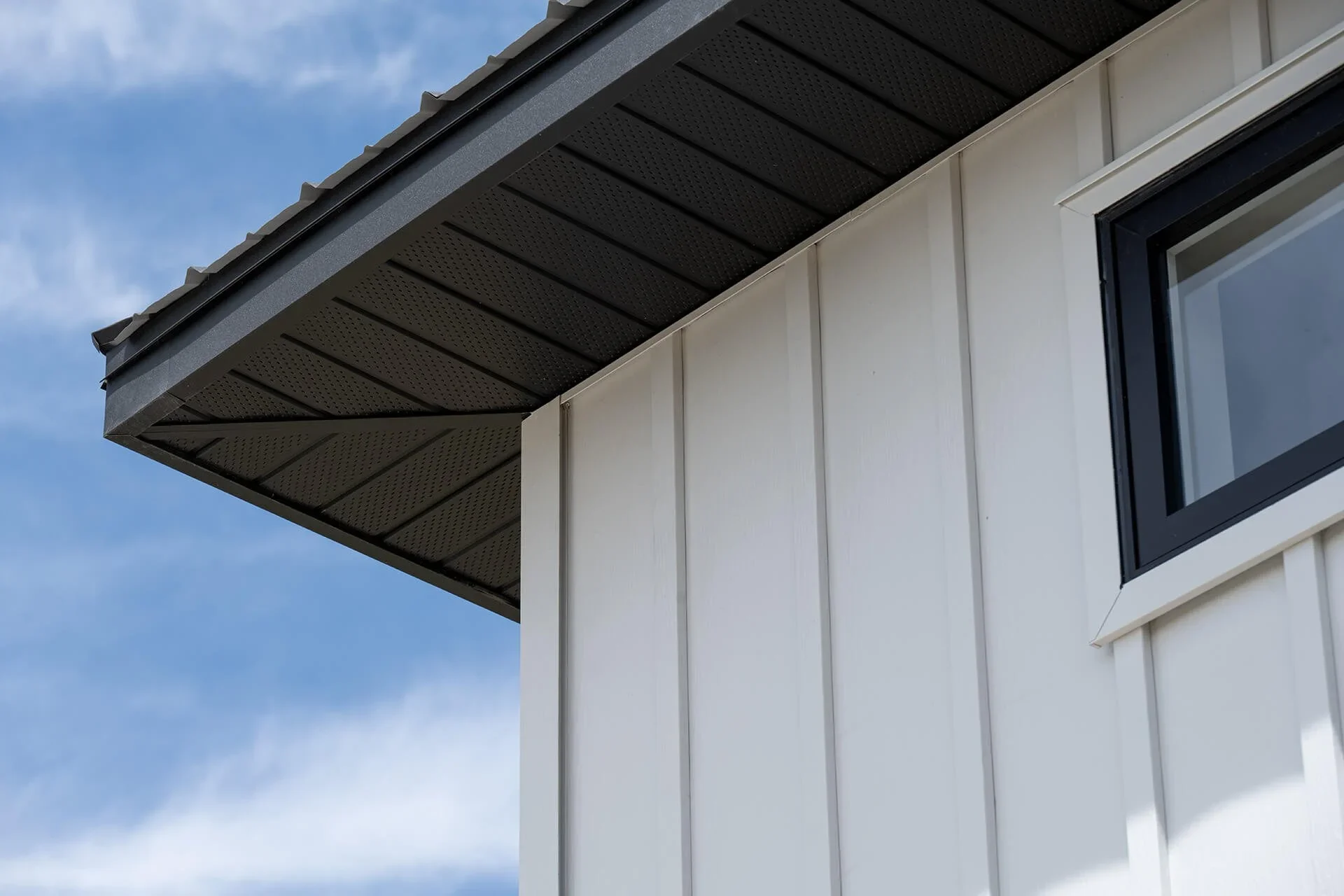 Close-up of the edge of a modern house's roof with dark roofing material and beige siding, a window with black trim, and a blue sky with some clouds in the background.