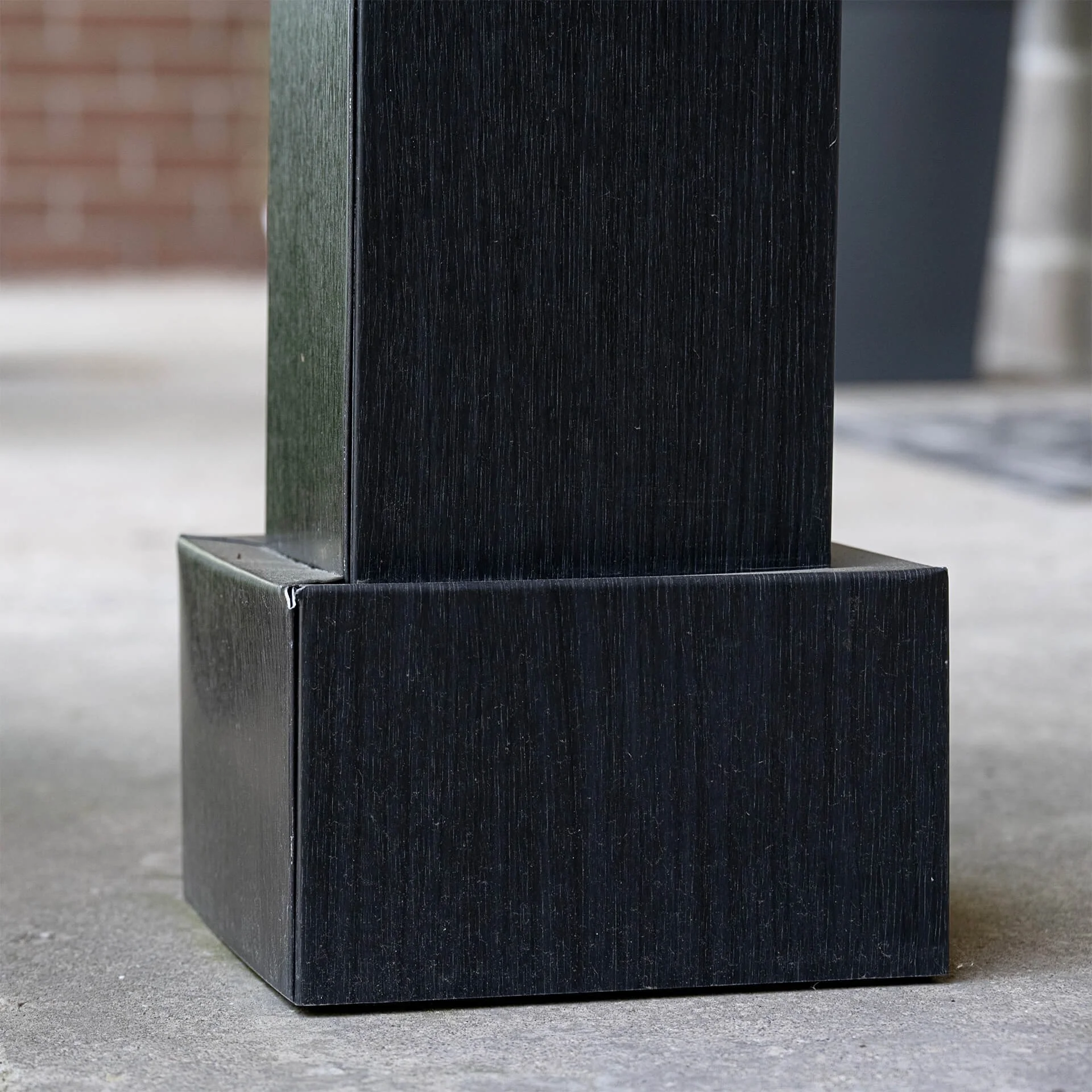 Close-up of a black, rectangular, wooden structure on a concrete surface, with a background showing blurred brick and gray elements.