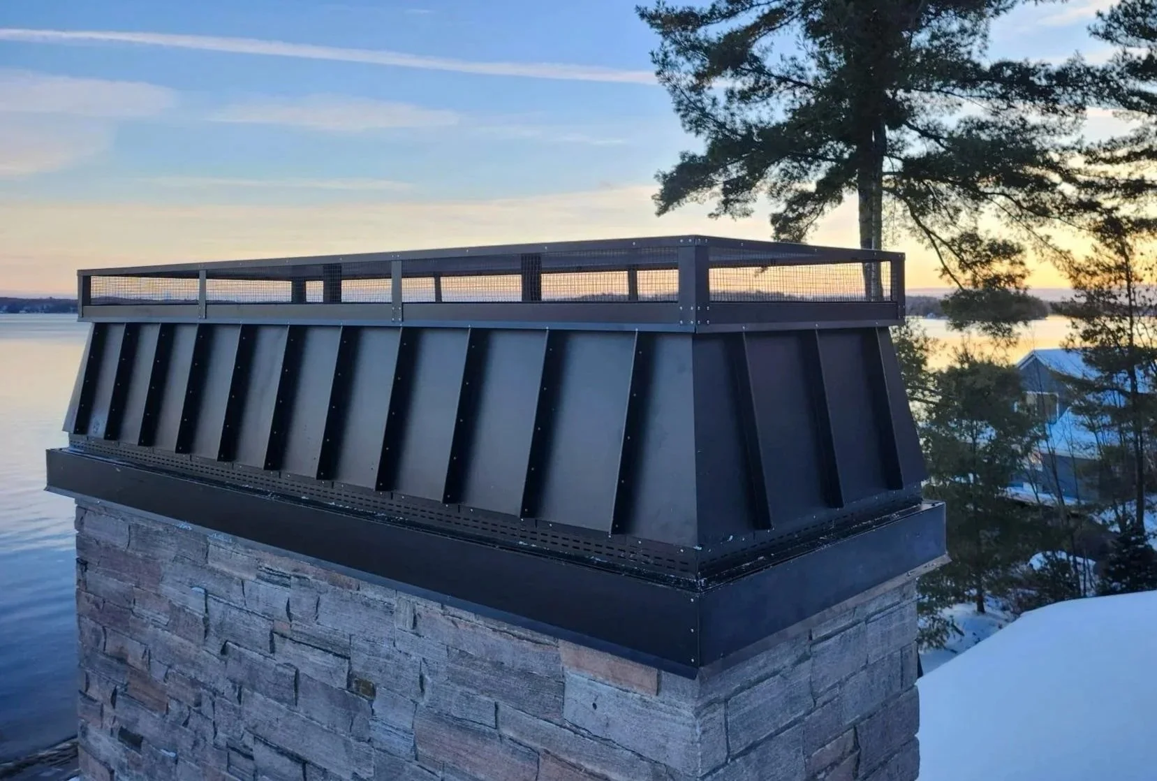 A metal chimney cap installed on a stone chimney on a lakeside during sunset, with trees and a building in the background.