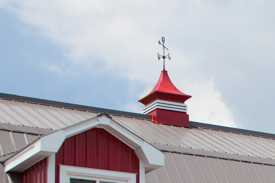 Close-up of a steeple with a weather vane on top of a red barn with a metal roof, under a cloudy sky.