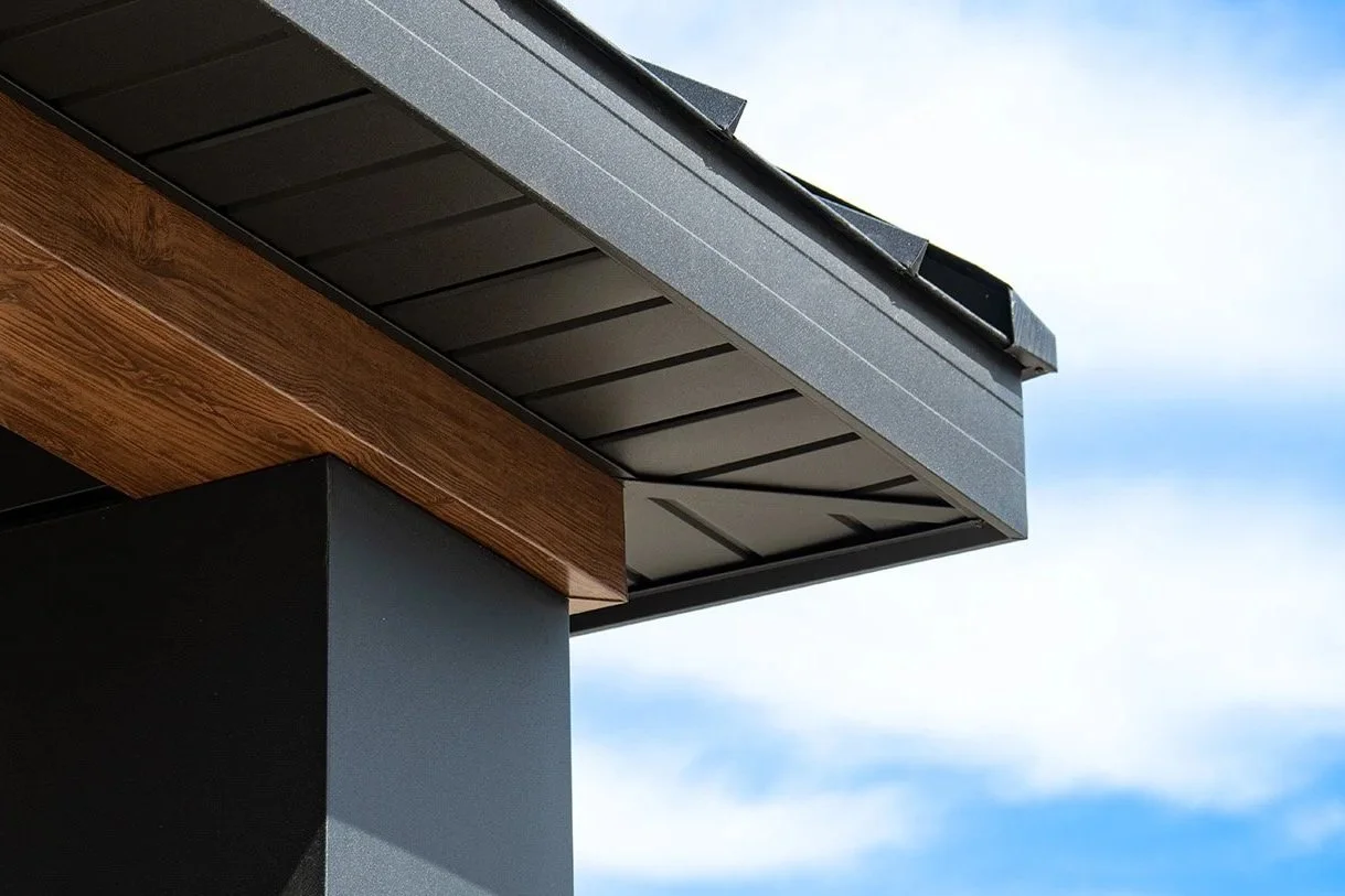 Close-up of a building corner showing a black roof overhang, a brown wooden trim, and black vertical wall against a blue sky with some clouds.