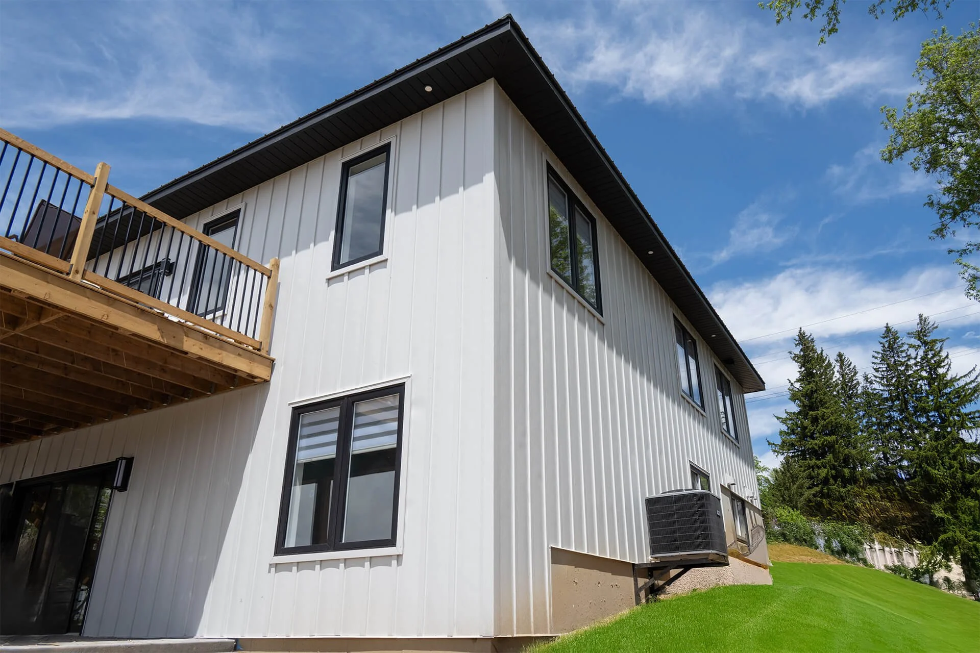 Exterior view of a modern, white, two-story house with black-framed windows, a wooden balcony, a black air conditioning unit, a green lawn, and tall trees under a partly cloudy sky.