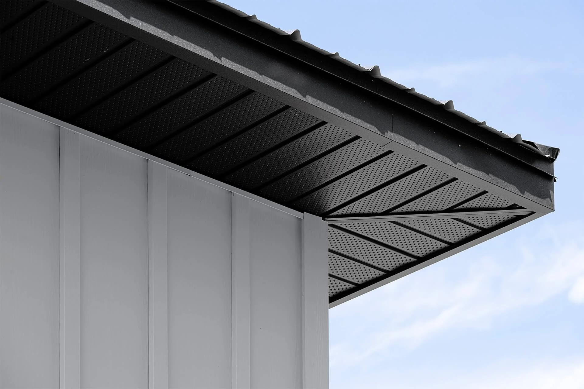 Close-up of the corner of a modern building's roof with black metal roofing and a white wall, with a patch of blue sky in the background.