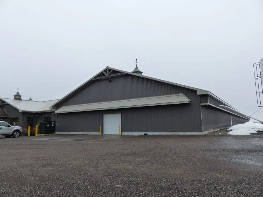 Large gray industrial or commercial building with a garage door, some snow on the ground, and a parking lot.