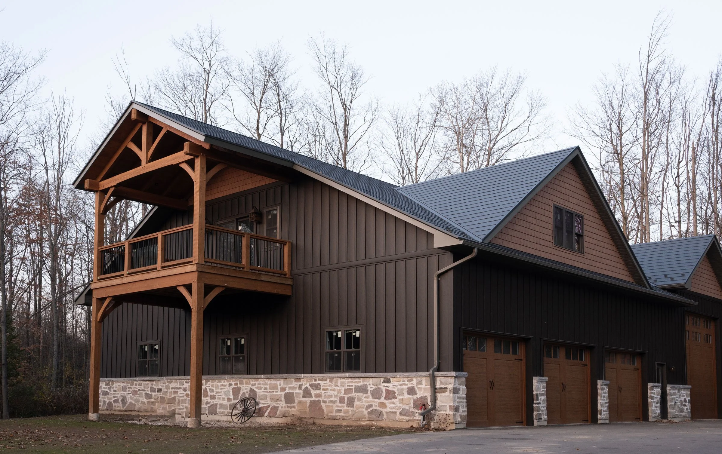 A modern residential house with dark vertical siding, a stone foundation, and wooden balcony and trim, set against leafless trees on a cloudy day.