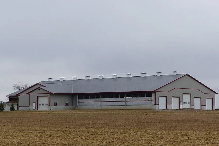 A large barn with gray walls and a gray roof, situated on a grassy field under an overcast sky.