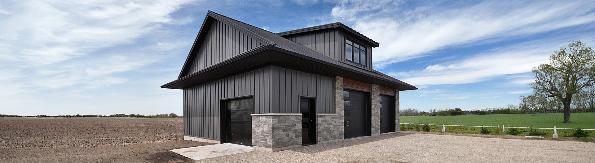 Two-story modern house with dark metal siding, stone accents, and large garage doors in a rural setting with open fields and a partly cloudy sky.