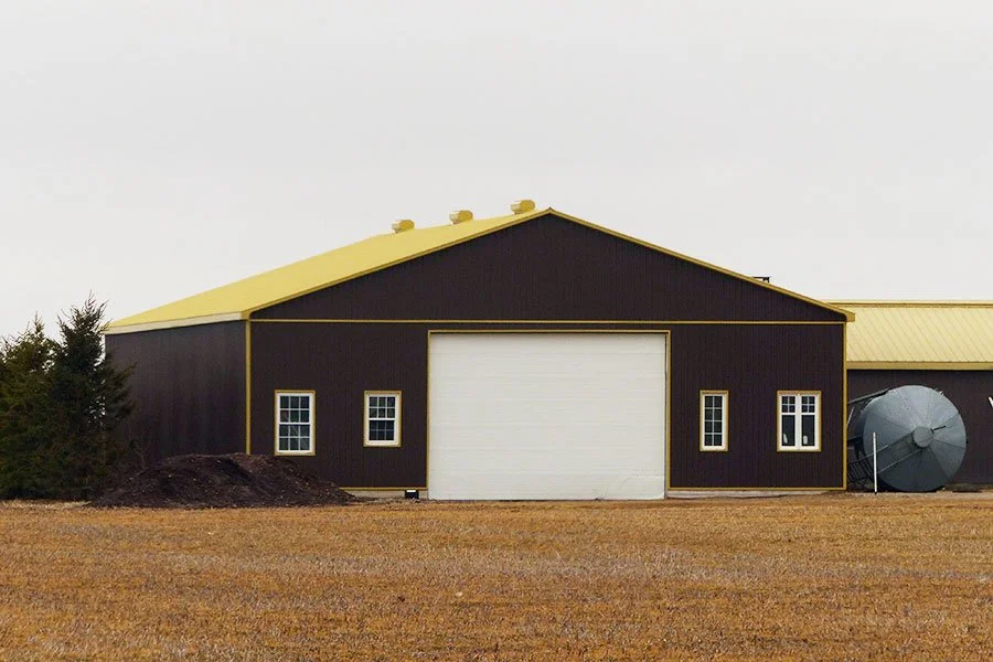 A large barn with dark brown siding, a yellow roof, and four windows, situated on a grassy field with some shrubs on the left and a satellite dish on the right.