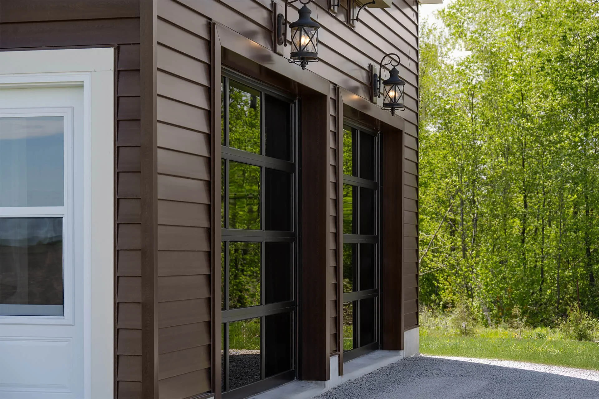 Exterior view of a modern house with brown siding, black framed glass doors, a white window, and two black outdoor lanterns on the wall, with green trees in the background.