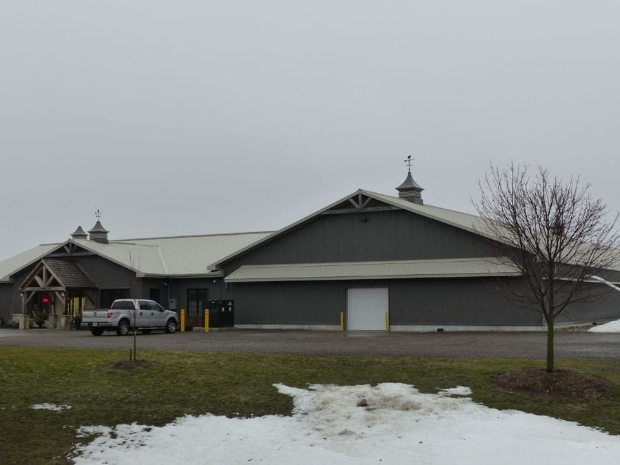 A large gray barn-style building with a metal roof, decorative cupolas, and a small clock tower, surrounded by a parking lot, leafless trees, and patches of snow on the ground.