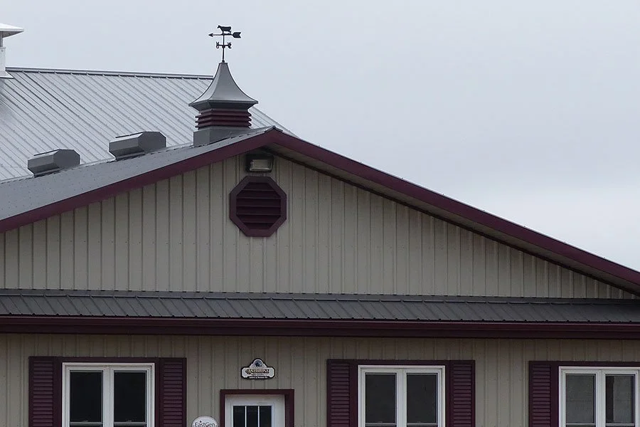 Close-up of the upper part of a beige building with purple trim, showing the roof, windows, and a small weather vane on top.