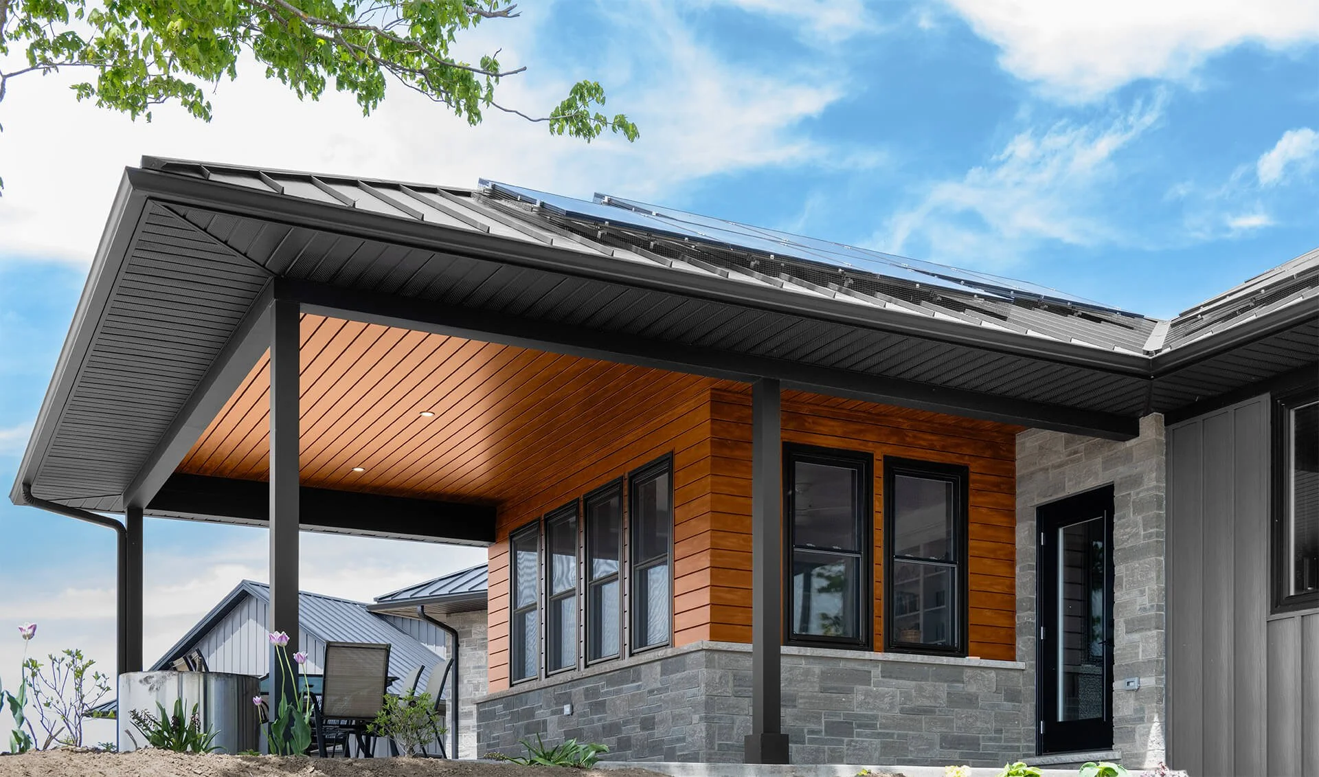 Modern house exterior with a covered patio, black window frames, gray brick and wood siding, and solar panels on the roof, against a blue sky with clouds.